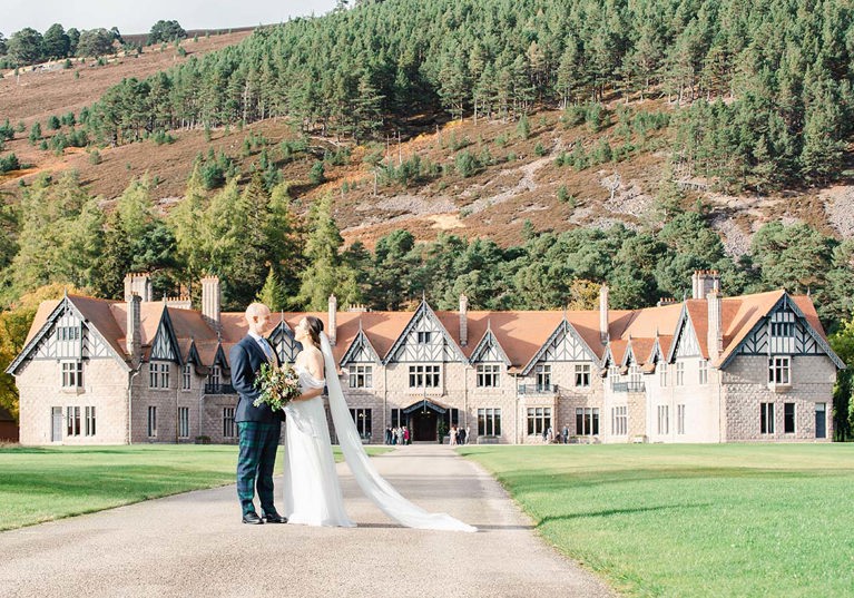 bride and groom standing in front of large country house with Scottish hills surrounding them